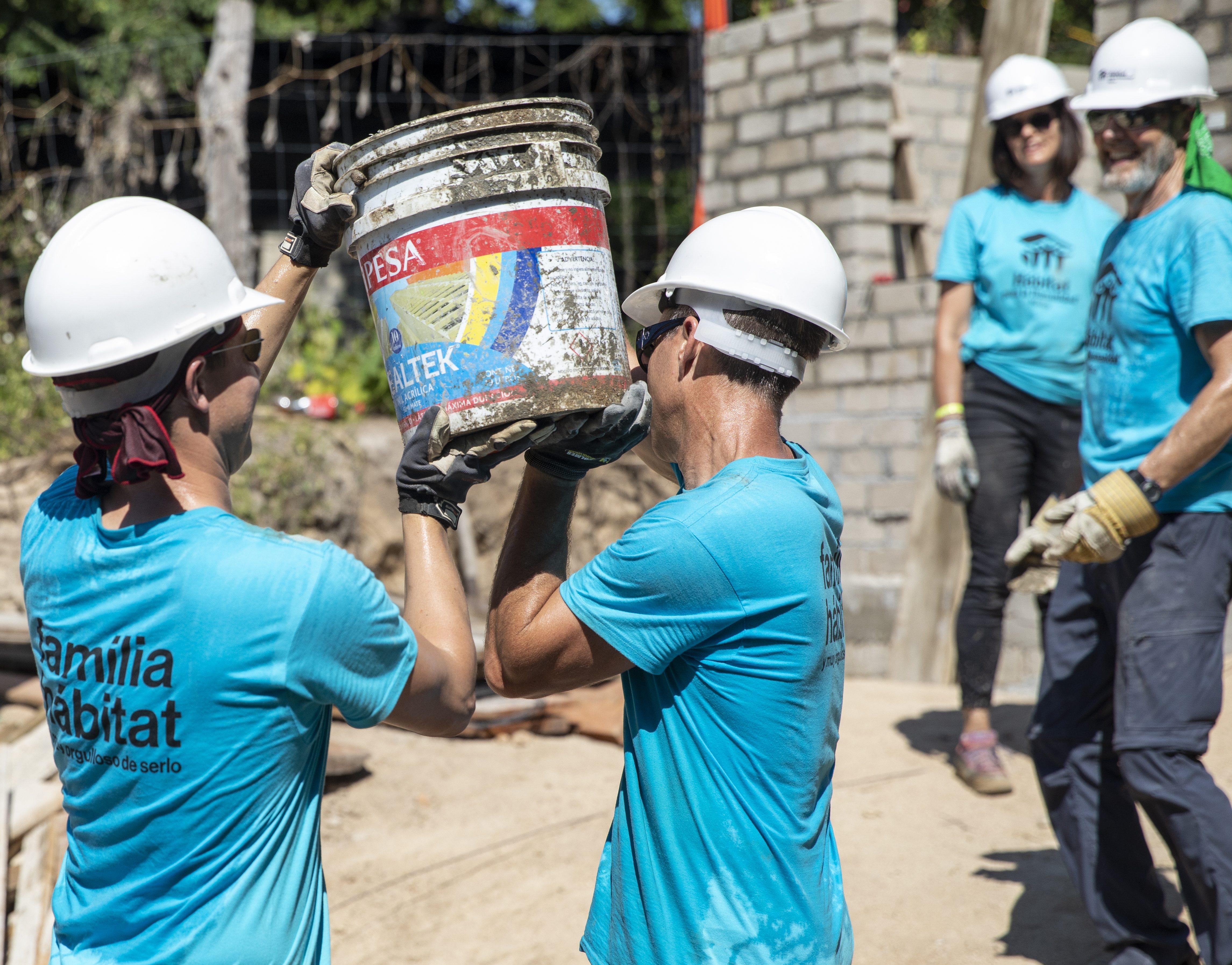 Two Habitat for Humanity Global Village volunteers carry a bucket of cement together.